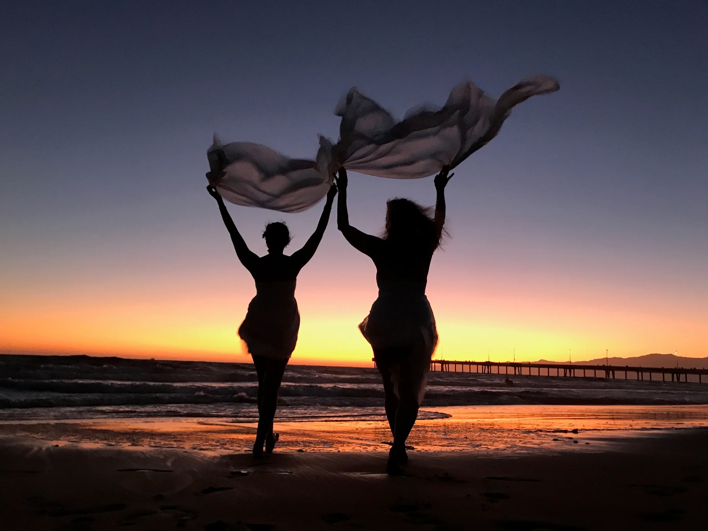 Two figures holding flowing fabric at an orange sunset